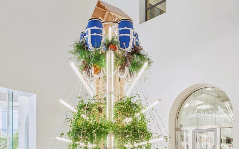 CON-VIVERE, The Edible Tower, is an architectural display that stands covered in natural materials like draping leaves and wooden frames in a light-filled gallery.